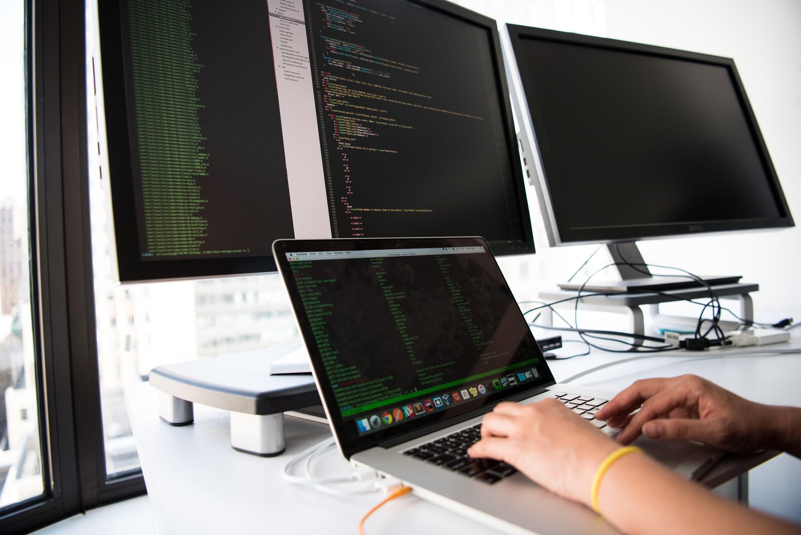Hände tippen auf einem MacBook; dahinter zwei Monitore, der linke zeigt Code in zwei Spalten, der rechte ist dunkel