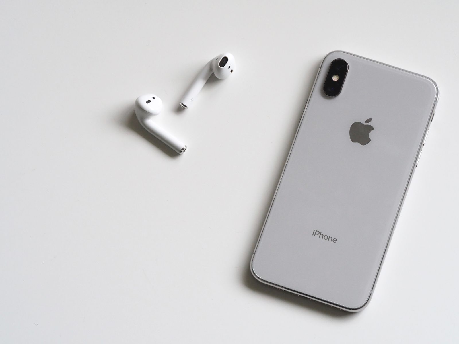 Ein silbernes iPhone liegt mit der Rückseite nach oben auf einem weißen Tisch, daneben ein Paar weiße AirPods