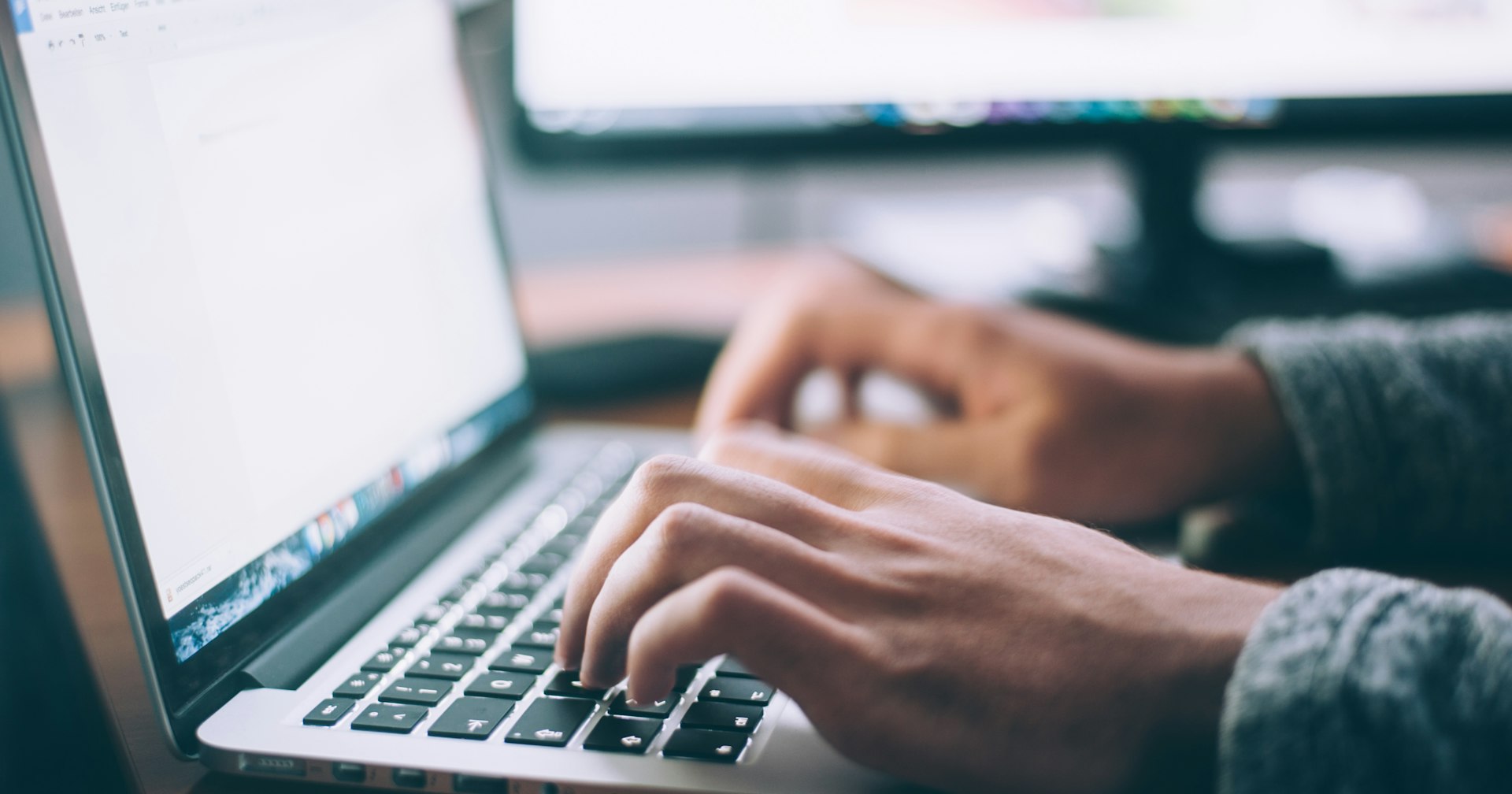 Hände tippen auf einem silbernen Laptop, unscharfer Monitor im Hintergrund