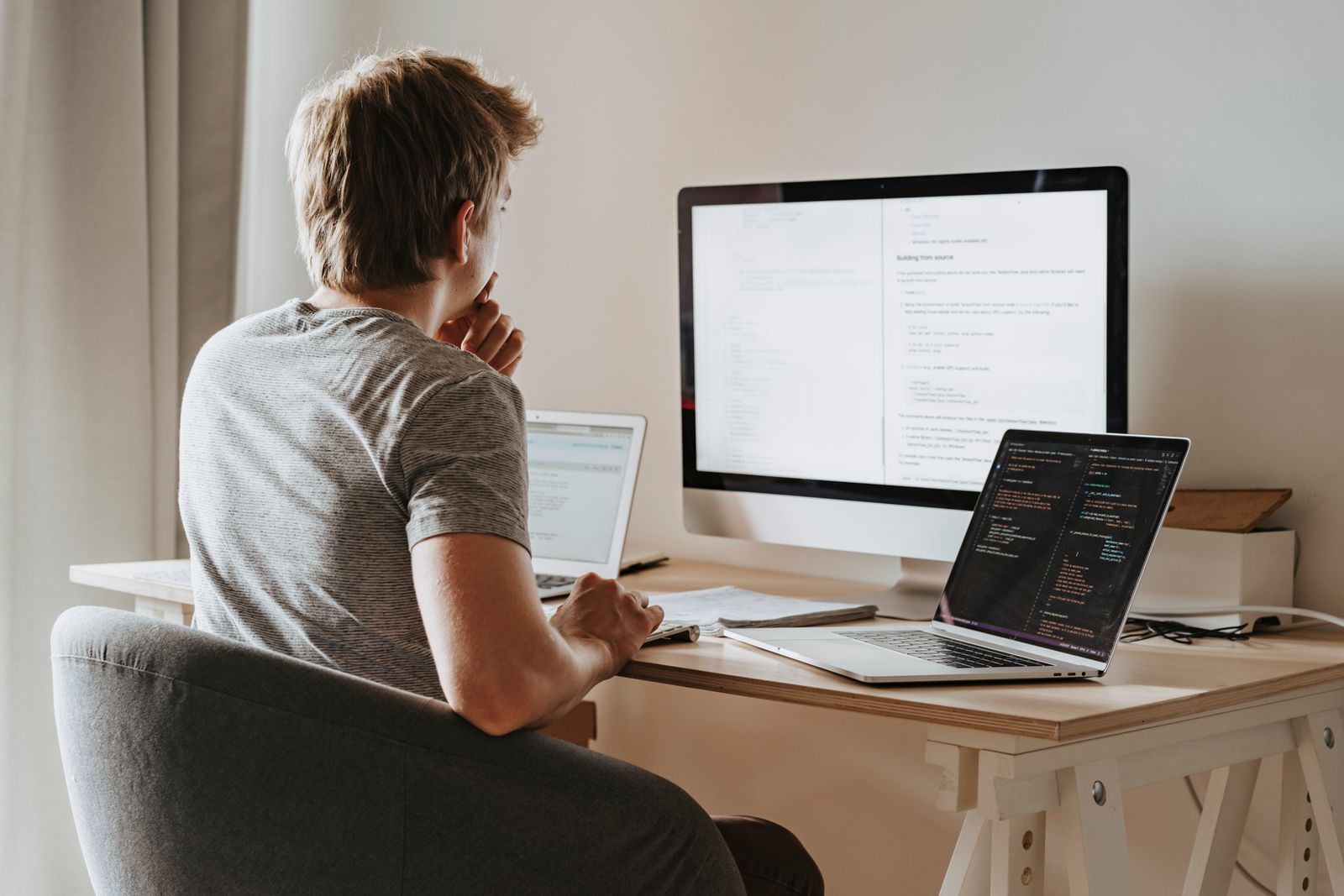 Une personne de dos est assise à un bureau en bois, avec un iMac, deux portables et des feuilles ; les écrans montrent du code et un texte