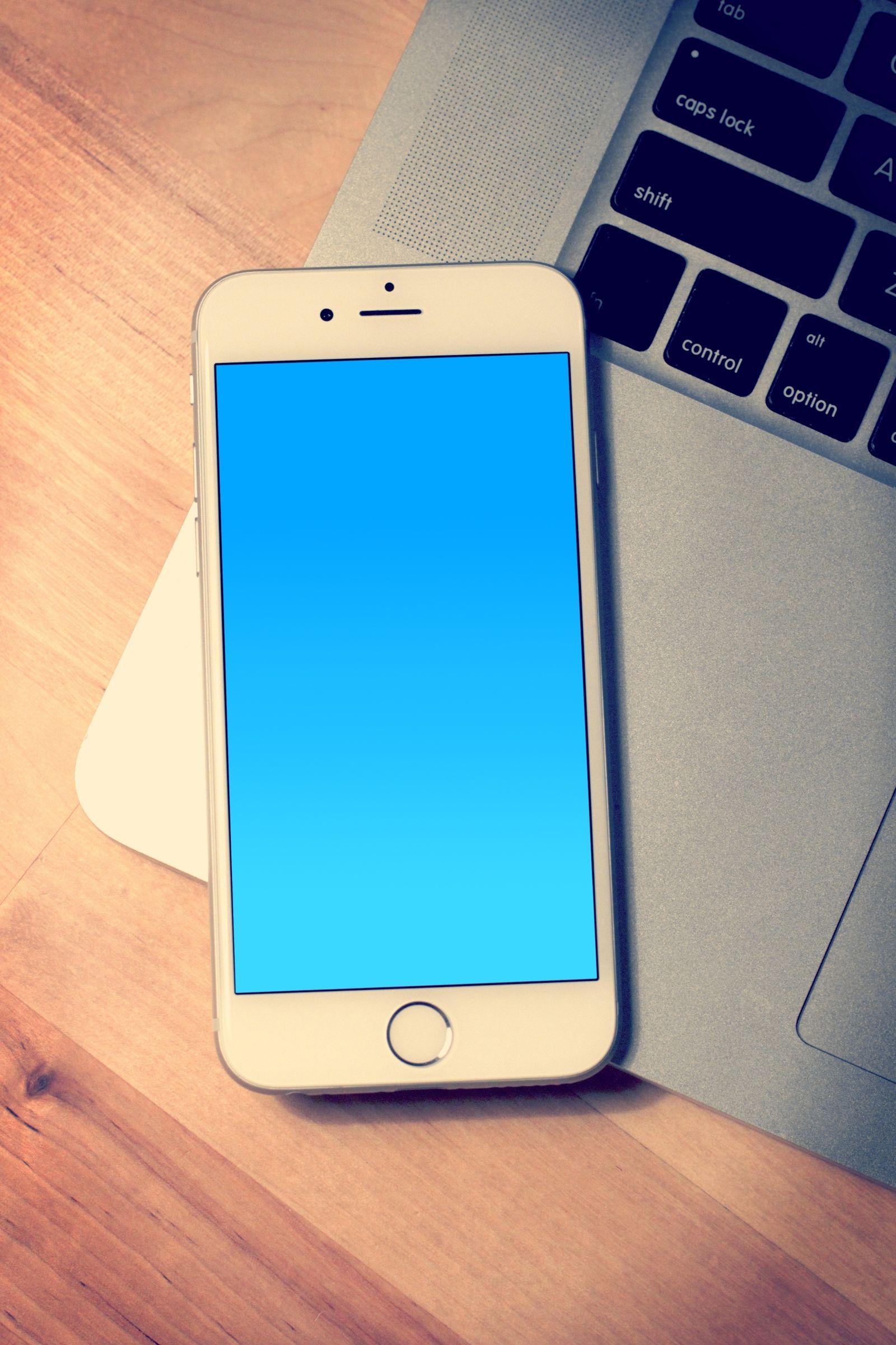 Un iPhone blanc à écran bleu uni est posé contre le clavier d’un MacBook argenté, sur une table en bois