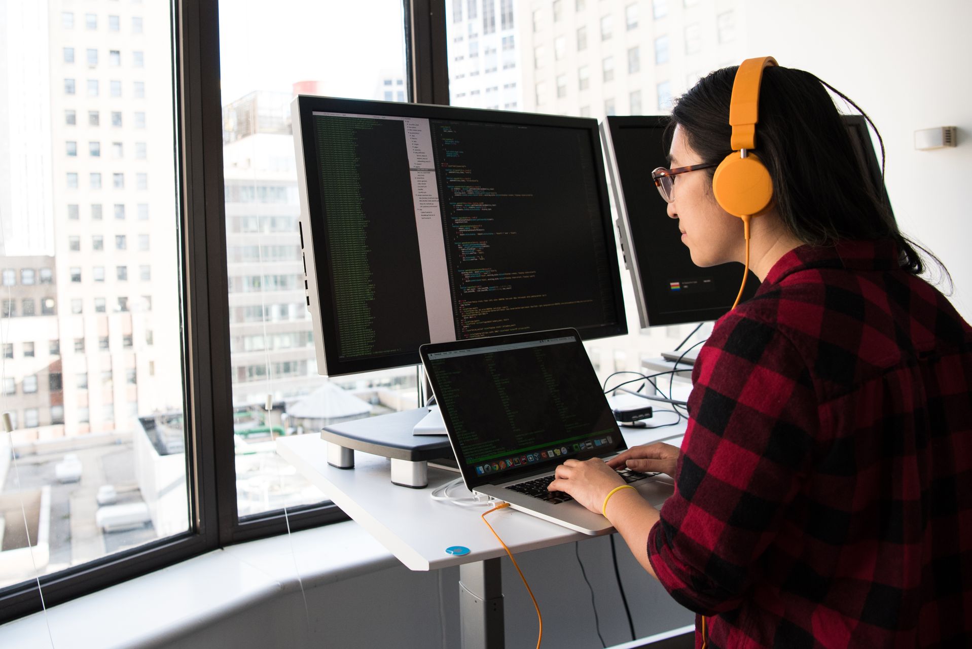 昇降デスク上の MacBook Pro と外部モニター二台、画面にコードエディタ