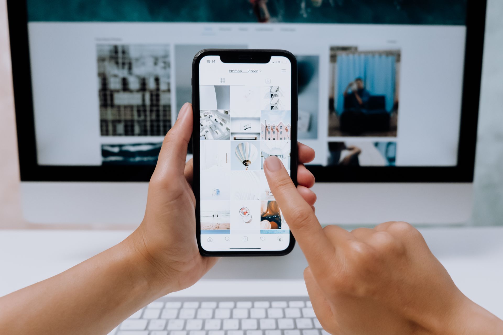 손 안의 iPhone과 뒤쪽 iMac·키보드가 보이는 책상